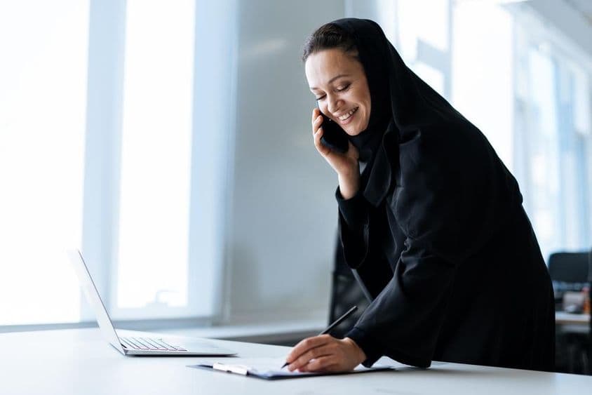 Una hermosa mujer con abaya trabajando en su computadora mientras está al teléfono.
