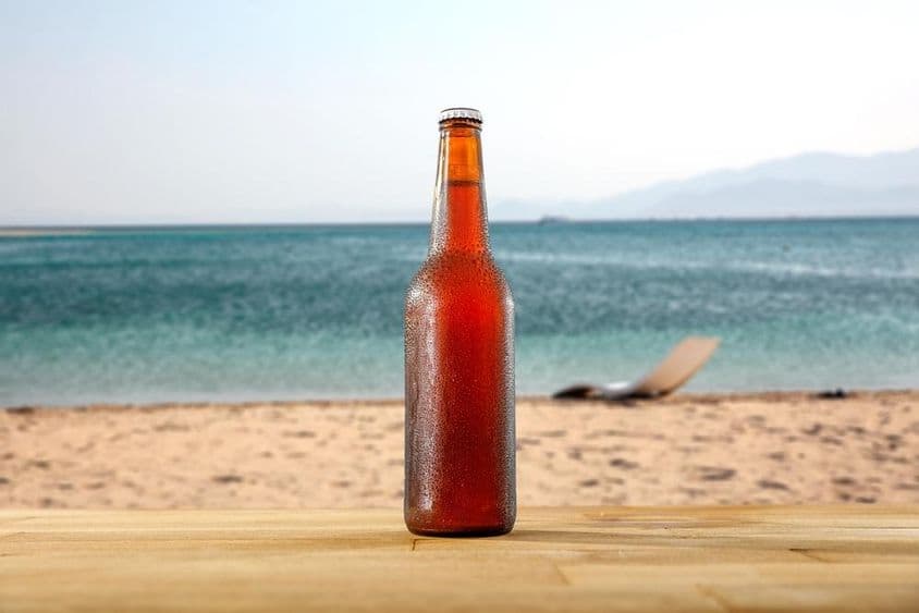 Una botella de cerveza fría sobre una mesa de madera en la orilla de una playa de arena.
