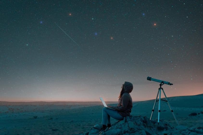 Mujer sentada en el desierto con una laptop junto a un telescopio en la noche, mirando el cielo estrellado.