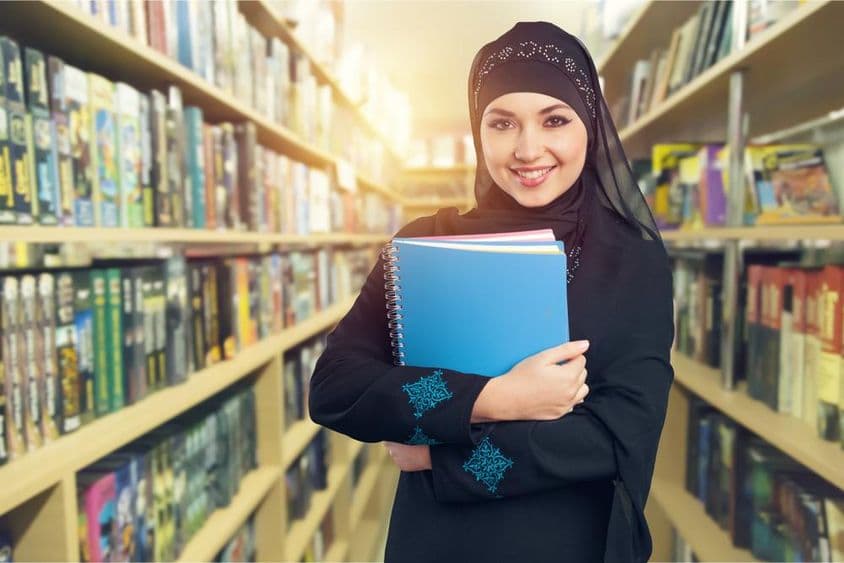 Estudiante árabe de pie en biblioteca con libros en el brazo.