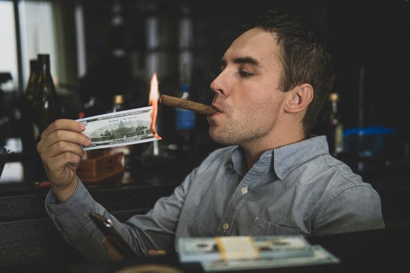 Joven apuesto encendiendo un cigarro con billete de cien dólares en un bar.