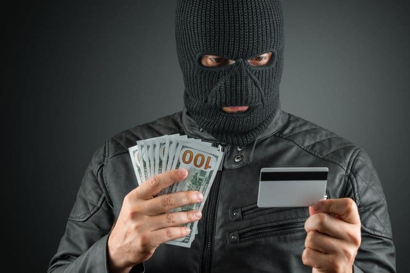 Criminal con pasamontañas sosteniendo tarjeta de crédito contra un fondo oscuro.