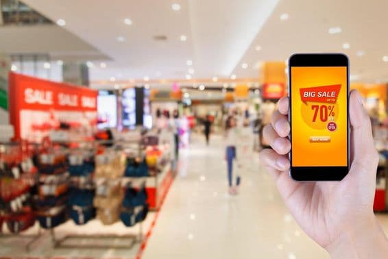 Smartphone mostrando pantalla de aplicación de gran venta con fondo de centro comercial desenfocado.