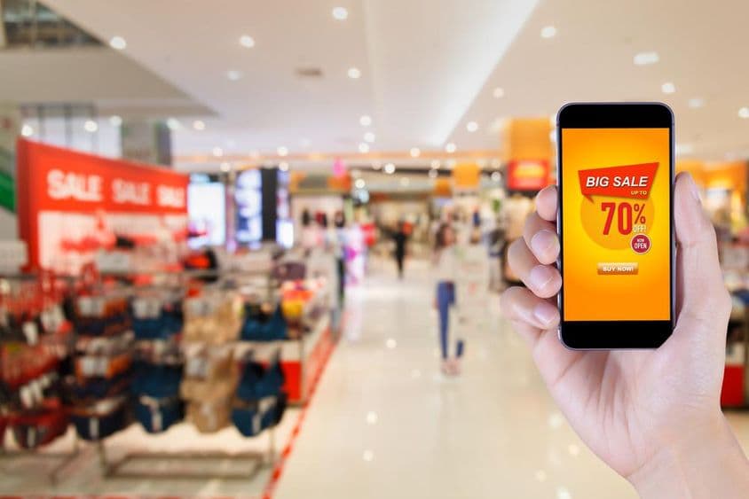 Smartphone mostrando pantalla de aplicación de gran venta con fondo de centro comercial desenfocado.