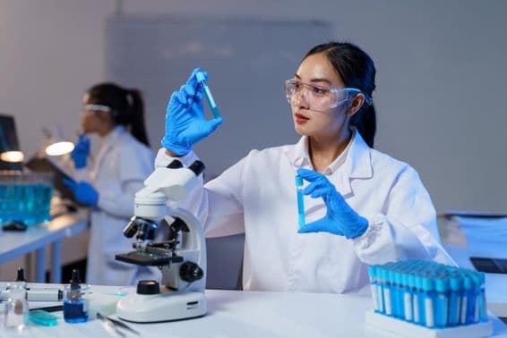 Científica asiática comparando dos tubos de ensayo en bata de laboratorio y guantes.