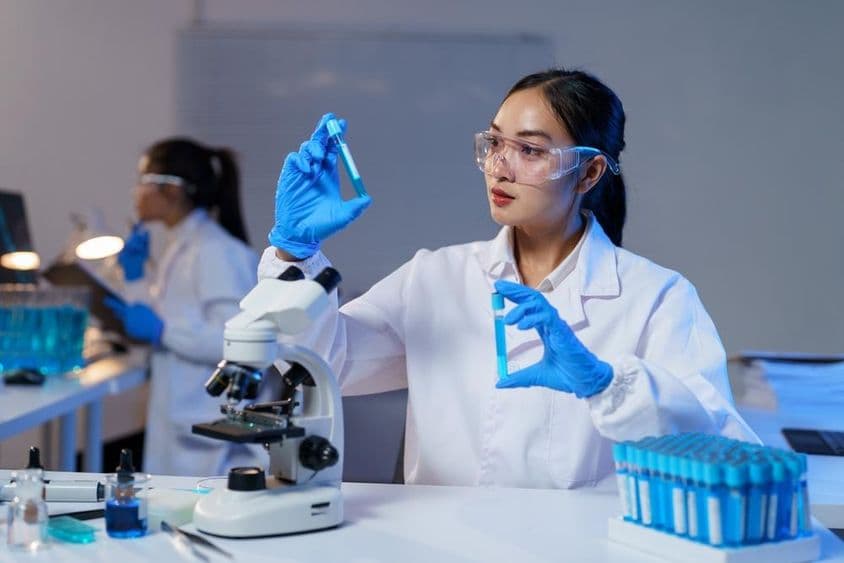 Científica asiática comparando dos tubos de ensayo en bata de laboratorio y guantes.