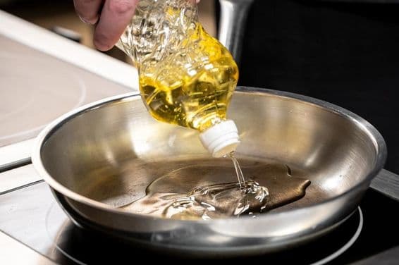 Una mano vertiendo aceite de cocina dorado desde una botella de plástico en un sartén de acero inoxidable.