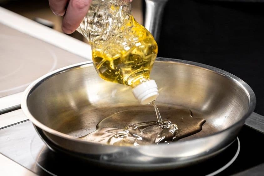 Una mano vertiendo aceite de cocina dorado desde una botella de plástico en un sartén de acero inoxidable.