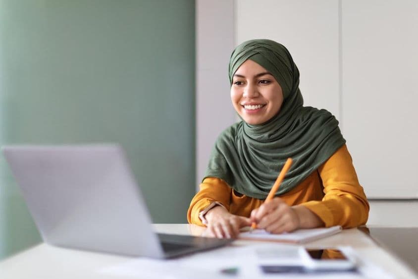 Joven mujer musulmana asistiendo a un webinar en casa en una laptop y tomando notas.