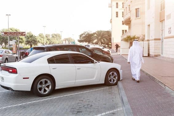 Un hombre con atuendo tradicional y un Dodge blanco en un estacionamiento.