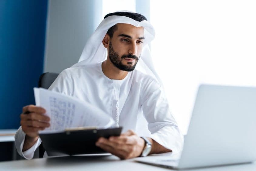 Hombre trabajando con una computadora portátil en una oficina de Dubái, vestindo traje tradicional blanco emiratí.