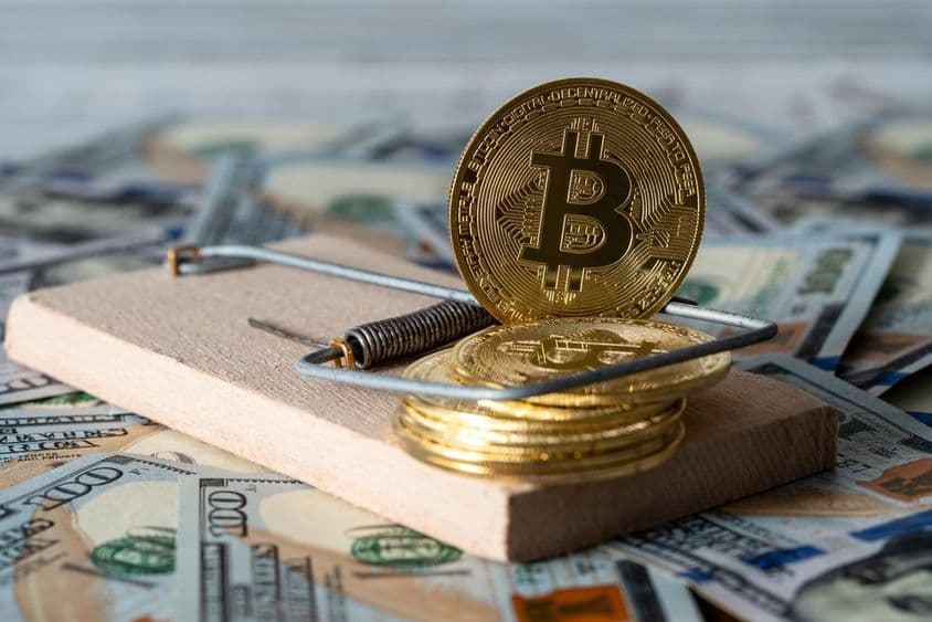 Monedas de criptomonedas doradas bitcoin en una trampa, con billetes de 100 dólares estadounidenses al fondo.