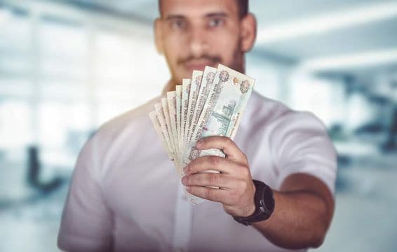 Hombre sosteniendo billetes de dirham, de pie frente a una sucursal bancaria.
