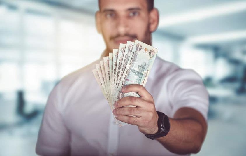 Hombre sosteniendo billetes de dirham, de pie frente a una sucursal bancaria.