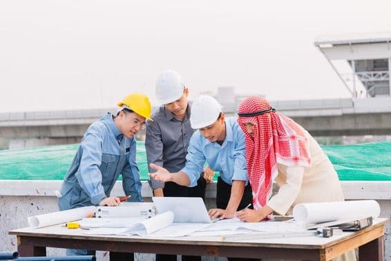 Empresario árabe, ingeniero y obrero interactúan con planos de construcción usando una laptop.