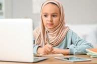 Niña musulmana pequeña con hijab haciendo tarea en casa.