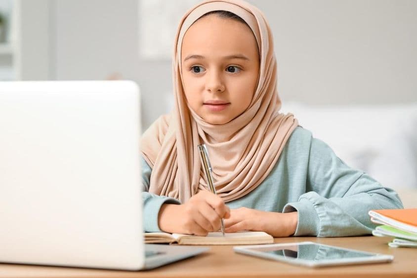 Niña musulmana pequeña con hijab haciendo tarea en casa.