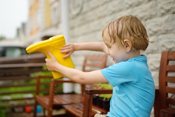 Niño preescolar empapado vertiendo agua de una bota amarilla de goma.