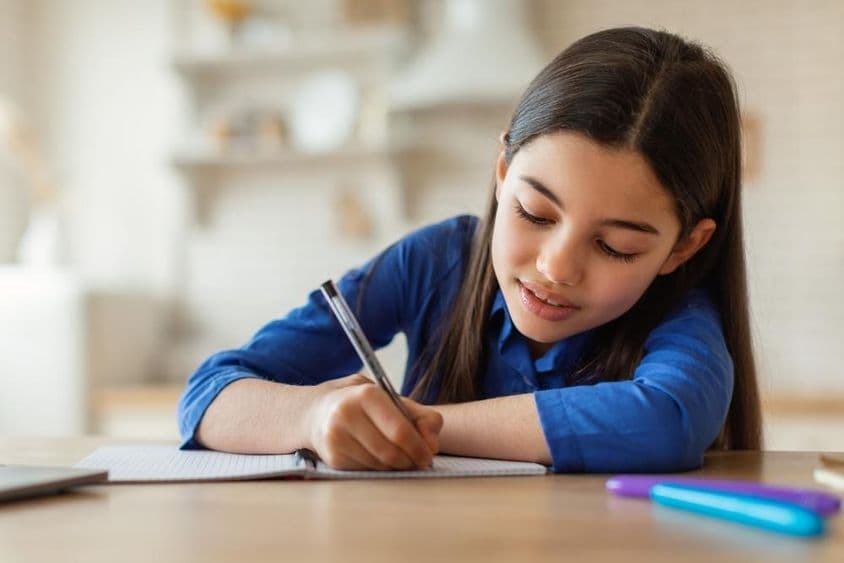 Niña árabe tomando apuntes mientras está sentada en una mesa con una laptop en casa.