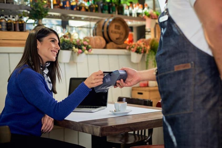 Mujer castaña sentada en una cafetería, pagando con tarjeta de crédito.