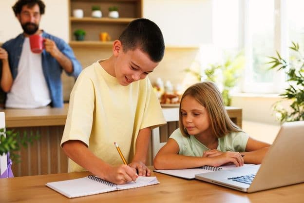 Hermanos haciendo tareas en casa usando computadoras portátiles y cuadernos.